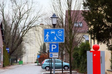 Sokak otoparkında ve yaya levhasında açık alanda mavi yol işareti var..