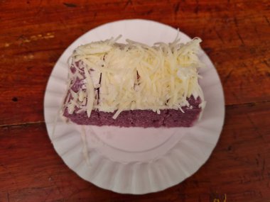 Ahşap bir masada beyaz bir kabın içinde peynirli, kasavadan yapılmış mor bir sandviç.