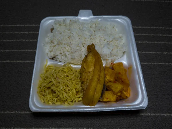 Food in a white container containing white rice, fried noodles, fried chicken and spicy fries