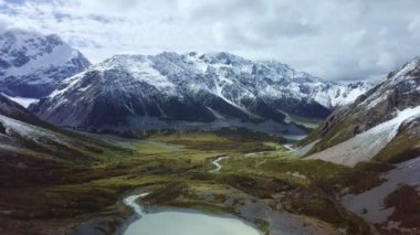 Buzul gölünden akan sığ akarsuyun hava görüntüsü ve karla kaplı sıradağların eteklerindeki yeşil vadi boyunca uzanır.