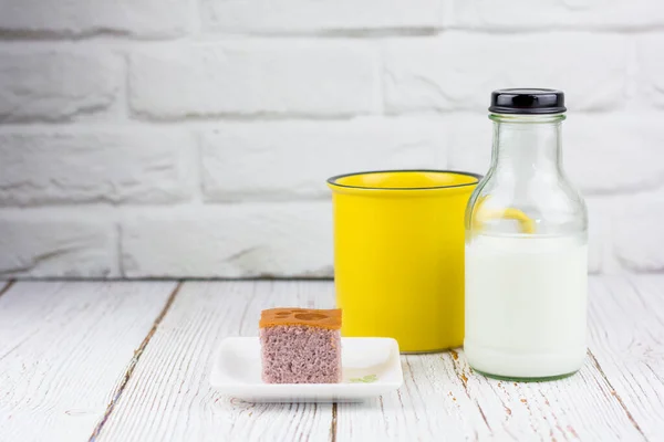 Yellow cup and a bottle of fresh milk served with a square-cut of sweet potato cake on a white plate in the kitchen. Bakery and beverage concept.