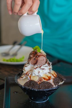 Çikolatalı dondurmanın üzerine sos dökmek, krema çırpmak, bingsu 'nun üzerine muz dilimi, siyah kâse, kafenin ve restoranın ahşap masasında bingsu yapmak. Lezzetli Kore buzlu dondurması.