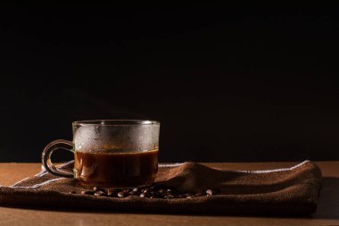 Bir fincan sıcak kahve dumanlı ve kahve çekirdekli kahve kahverengi masa örtüsü üzerine siyah arkaplan ve metin için fotokopi alanı. Kahve konseptinin faydaları.