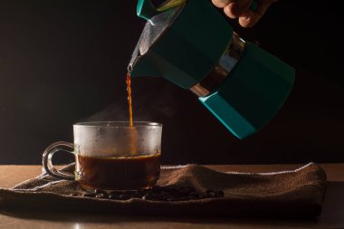 Kahverengi masa örtüsü ve ahşap masa üzerinde kahve çekirdeği ve dumanlı kahve fincanını temizlemek için yeşil moka demliğinden sade kahve dök. Kahve konseptinin faydaları.