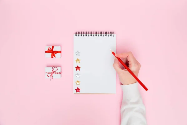 Christmas cozy background. Female hand writing in blank notebook with white gift boxes with red bows on pastel pink. New year planning, goals, to-do list or wish list concept. Flat lay, copy space. — Foto Stock