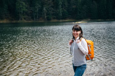 Ormanla çevrili dağ gölünün kıyısında duran turuncu sırt çantalı mutlu genç kız. Seyahat kavramı.