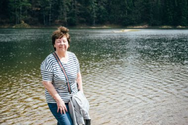 Dağ gölünün kıyısında dikilen yaşlı, gülümseyen bir kadın. Gözleri orman tarafından kapatılmış. Seyahat kavramı.