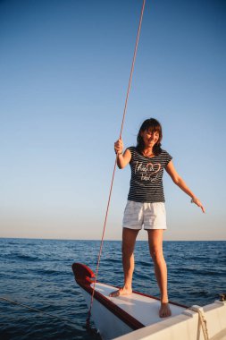 Young pretty smiling woman in striped shirt and white shorts at luxury yacht in sea, sunset.