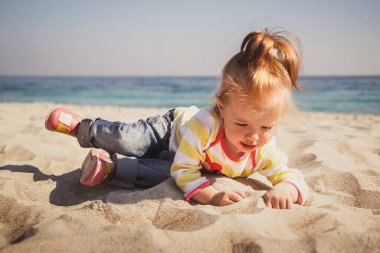 Küçük bebek, kot pantolonlu küçük kız, pembe ayakkabılar ve renkli kazak kumsalda oturuyor ve kumsalda oynuyor..