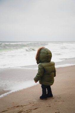 Sıcak ceket giyen küçük yalnız bir çocuk okyanus kıyısındaki fırtınaya bakıyor..