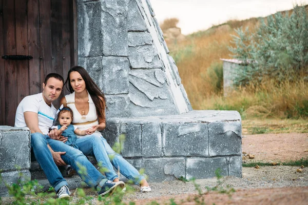 Happy young family in white t-shirts and blue jeans with a small daughter in dress sitting near to the lighthouse.
