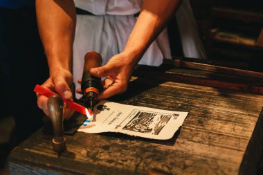Bled, Slovenia - September, 8 2018: Man's hands making souvenir, melting red sealing wax candle with burner for personal stamp at old-style picture post card. Souvenir shop at Bled Castle, Slovenia.