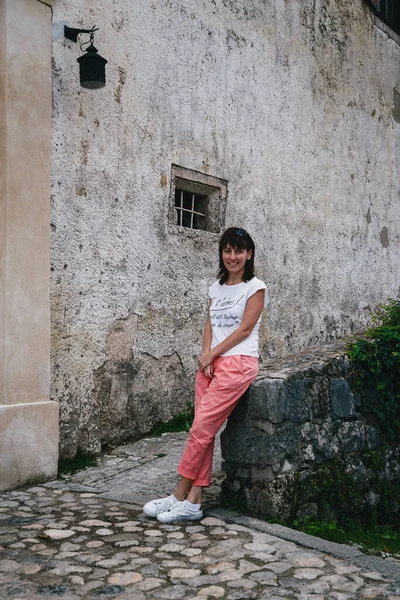 Young smiling woman tourist sitting on a Bled Castle wall in the courtyard. Wearing white shirt, white sneakers and coral trousers. Travel concept