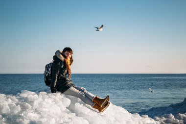 Young smiling woman sitting on a top of frozen sea ice blocks on a coast of the sea with a blue sky at the background, at frosty sunny day
