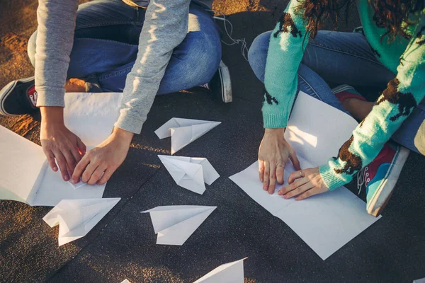 Teenagers making paper airplanes sitting at the roof of a building at sunset. Young people holding paper aeroplanes, love, relations concept.