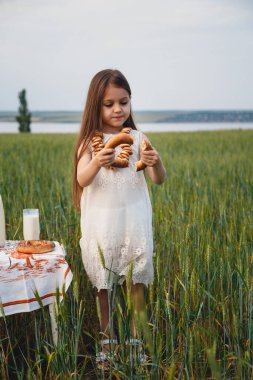 Cute little girl in white dress eating bagels with milk in green field.