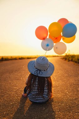 Little girl sitting on road with colorful balloons in hands at sunset. Back view.