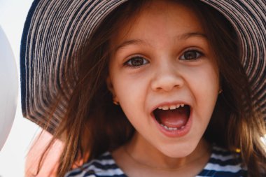 Close up fun portrait of cute laughing little girl in striped sunhat and shirt looking at camera. Selective focus.
