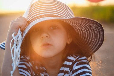 Little serious girl sitting on road with colorful balloons in hands at sunset.