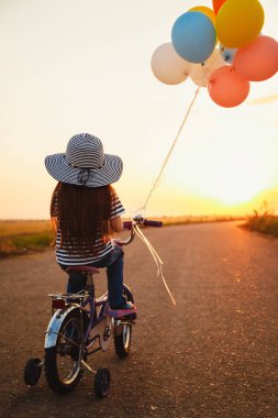 Cute little girl with colorful balloons cycling at the road at sunset sky summer time. Back view