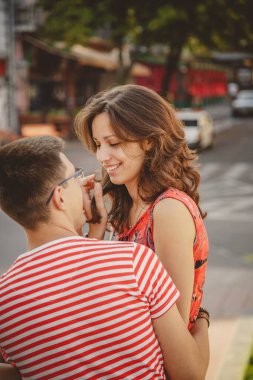 Cute young smiling couple in love laughing, hugging, sitting outdoors at green city street, looking at each other, summertime.