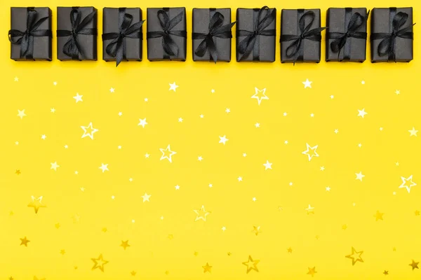 Black gift boxes with black ribbons and golden glitter stars confetti sparkles on bright rich yellow background. Black Friday sale flat lay, top view, copy space. Holiday, festive, party backdrop