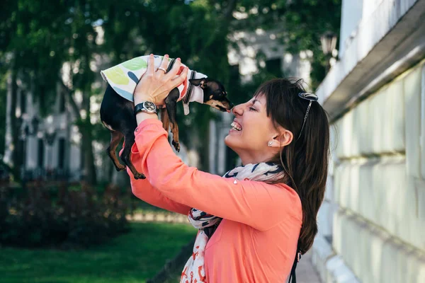 Happy young caucasian woman holding her small dog which kissing her face. Russian Toy or Russian Toy Terrier, dressed in autumn suit. Love pets concept