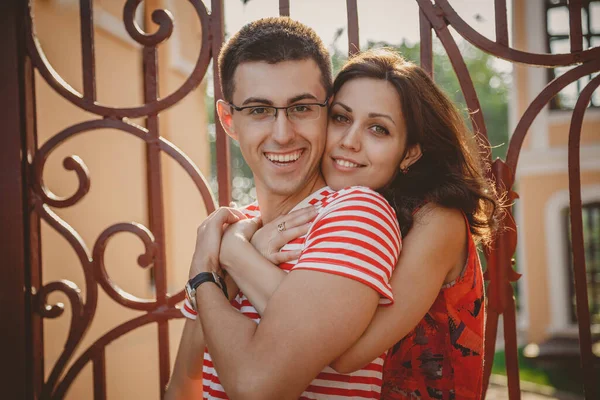 Close up of beautiful young smiling couple in love, hugging, standing behind each other outdoors at city street. Looking at camera.