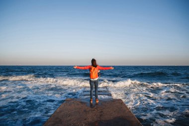 Turuncu sırt çantalı genç mutlu kız deniz kenarında duruyor temiz hava soluyor ve kollarını kaldırıyor manzaranın tadını çıkarıyor..