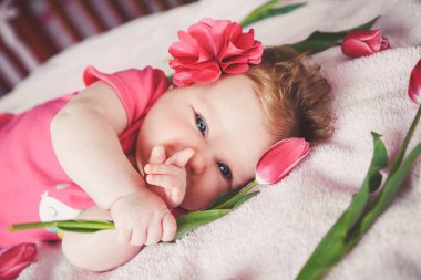 Close-up portrait of cute smiling baby girl lying down on pink bed with tulip in hand. Looking at camera and pointing finger