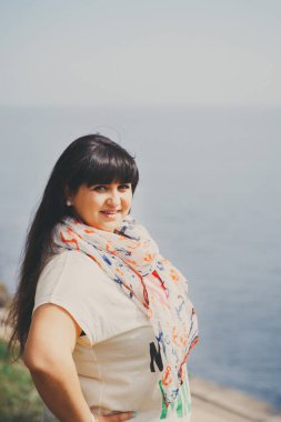 Mutlu gülümseyen, şişman, beyaz tişörtlü, atkısı açık denize yakın genç bir kadın. Kendine güvenen şişman genç kadın. XXI kadın, artı doğadaki boy kadın.