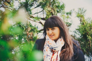 Koyu mavi ceketli ve açık havada çapalı atkısı olan mutlu gülümseyen şişman bir kadın. Kendine güvenen şişman genç kadın. XXI kadın, artı doğadaki boy kadın.