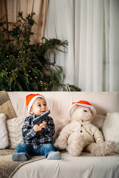 Christmas portrait of happy smiling little boy in red santa hat sitting on sofa playing with toy gun present. Winter holiday Xmas and New Year concept.
