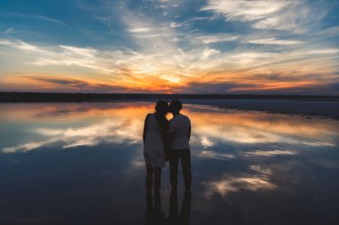 Portrait, silhouette of happy couple watching the colourful bright sunset standing in large lake and kissing, reflection in the water, holding hands.