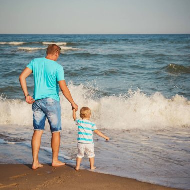 Young father with small son walking at beach together near the ocean, happy lifestyle family concept. Back view.