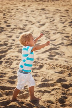 Happy small stylish boy in white shorts and striped blue t-shirt enjoying life on summer beach.