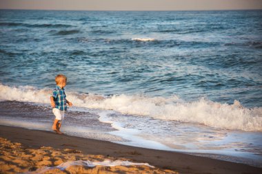 Happy small stylish boy in white shorts and blue shirt enjoying life on summer beach.