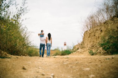 Back view of young family with a small kid walking to the white lighthouse, outdoors background.