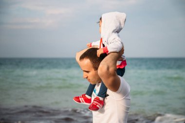 Close up of small baby girl in hood sitting on father's shoulders on the sky background.