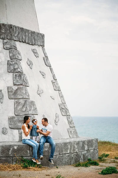 Happy young family in white t-shirts and blue jeans with a small daughter in dress near to the lighthouse, playing together.