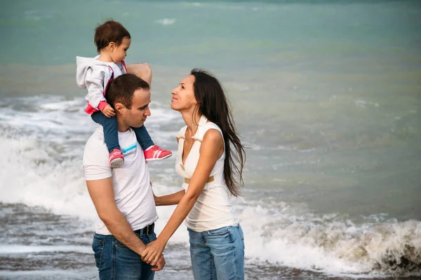 Happy young family in white t-shirts and blue jeans with a small daughter walking along the seaside.