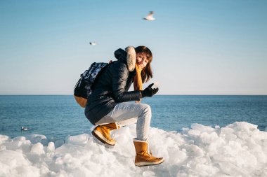 Young smiling woman sitting on the top of sea ice blocks on the coast holding broken piece of ice, with a blue sea and sky and the background