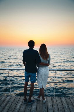Young couple watching the sunrise at the beach, back view, seashore summer beach at yellow blue morning horizon sea.