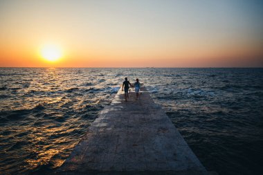 Young couple running at the pier towards the sunrise sun at the beach summer time.