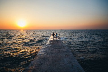 Young couple running at the pier towards the sunrise sun at the beach summer time.