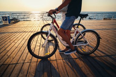 Close up of couple of young hipsters cycling together at the beach at sunrise sky at wooden deck summer time.