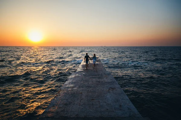 Young couple running at the pier towards the sunrise sun at the beach summer time.