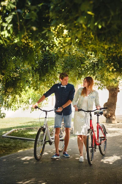 Happy stylish loving couple walking with bicycles in a park.