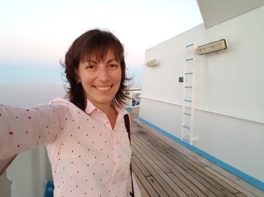 Young smiling woman in soft pink shirt making selfie photo on board of cruise ship at open deck to share on internet social media