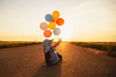 Little girl sitting on road and playing with colorful balloons at sunset. Back view.